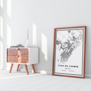 May include: A framed black and white map of Playa del Carmen, Mexico, with the city name and coordinates. The map is in a brown wooden frame, leaning against a white wall. A white and wood-tone nightstand with a vase and books is to the left.