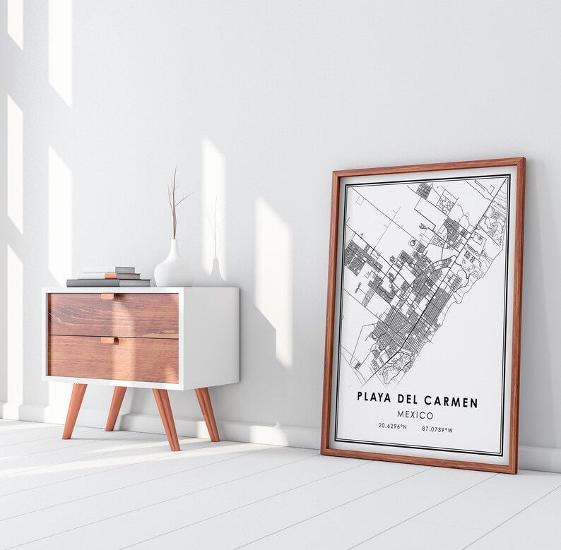 May include: A framed black and white map of Playa del Carmen, Mexico, with the city name and coordinates. The map is in a brown wooden frame, leaning against a white wall. A white and wood-tone nightstand with a vase and books is to the left.