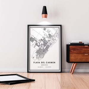 May include: A framed black and white map print of Playa del Carmen, Mexico, with the city's coordinates. The print is displayed on a wooden floor, leaning against a white wall. A black frame lies on the floor in front of the print.