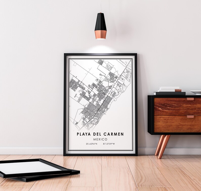 May include: A framed black and white map print of Playa del Carmen, Mexico, with the city's coordinates. The print is displayed on a wooden floor, leaning against a white wall. A black frame lies on the floor in front of the print.