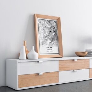May include: A framed black and white map of Punjab, Pakistan, with the text "Punjab, Pakistan 31.1784&deg;N 72.9879&deg;E" printed below the map. The frame is light brown wood and the map is sitting on a white and light brown dresser with six drawers.