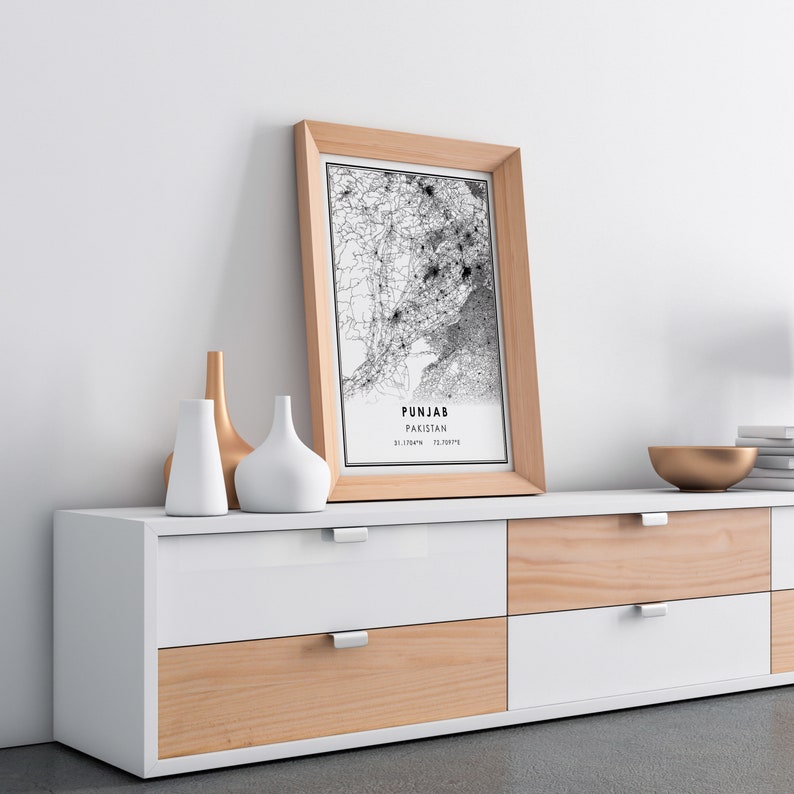 May include: A framed black and white map of Punjab, Pakistan, with the text "Punjab, Pakistan 31.1784&deg;N 72.9879&deg;E" printed below the map. The frame is light brown wood and the map is sitting on a white and light brown dresser with six drawers.