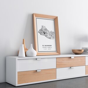 May include: A framed black and white map of El Salvador, Central America, with the coordinates 13.7947&deg; N, 88.8965&deg; W. The map is hanging on a white wall above a white and wood dresser with six drawers.