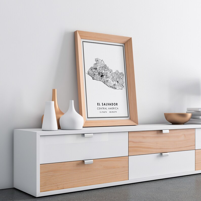 May include: A framed black and white map of El Salvador, Central America, with the coordinates 13.7947&deg; N, 88.8965&deg; W. The map is hanging on a white wall above a white and wood dresser with six drawers.