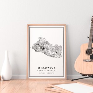 May include: A framed black and white map of El Salvador, Central America with the coordinates 13.7942&deg;N 88.8963&deg;W printed below the map. The map is hanging on a white wall with a wooden floor and a brown acoustic guitar in the background.