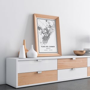May include: A framed map print of Playa del Carmen, Mexico, in a light wood frame. The print is black and white, with the city's layout detailed. The frame rests on a white and wood-toned cabinet, alongside decorative vases and a gold bowl.