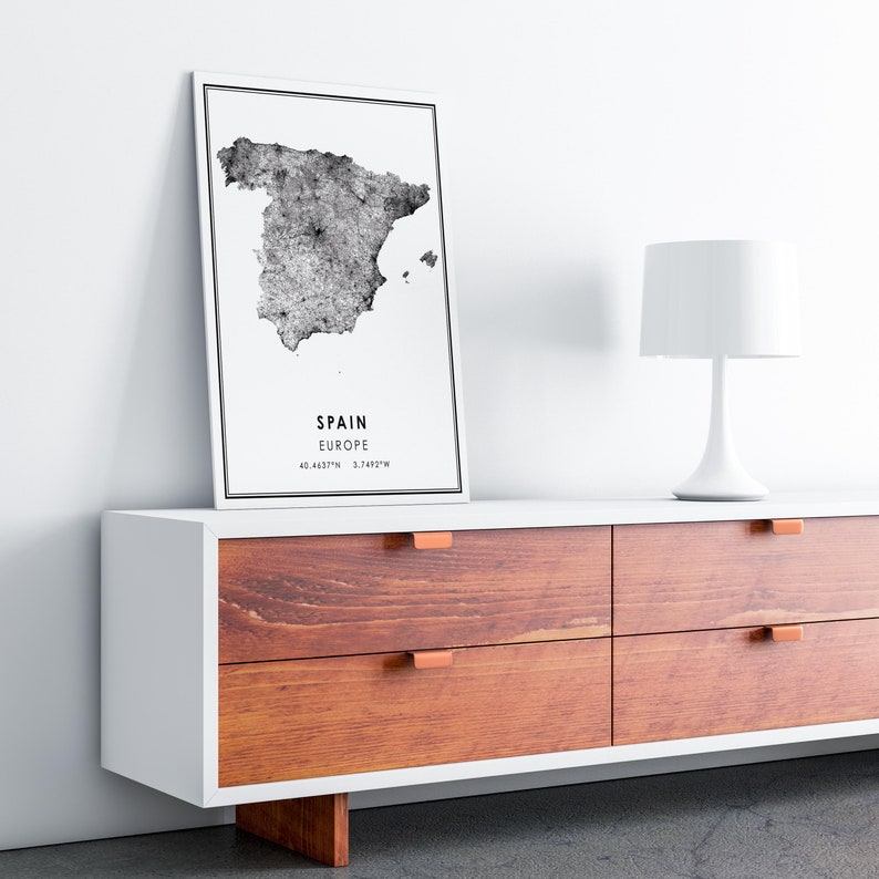 May include: A black and white map print of Spain with the text "SPAIN EUROPE 40.4637&deg;N 3.7492&deg;W" on a white background. The print is hanging on a white wall above a white dresser with four drawers. The dresser has a wood grain finish and copper drawer pulls. A white table lamp is on the dresser.