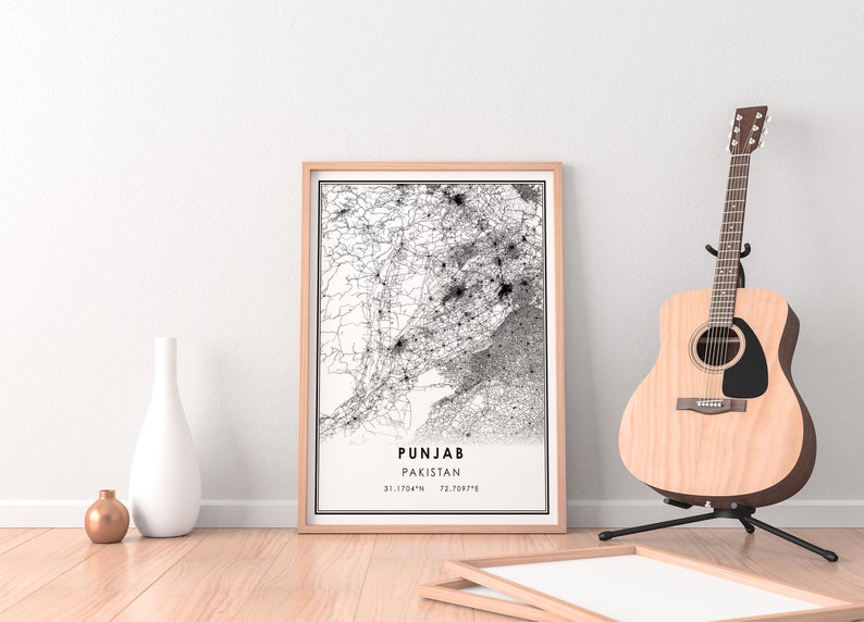 May include: A black and white map print of Punjab, Pakistan, in a light wood frame. The map shows the roads and cities of the region. The print is hanging on a white wall with a wooden floor and a guitar in the background.