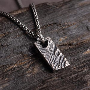 May include: A rectangular silver pendant with a textured, rugged surface and a dark oxidized finish. The pendant hangs from a silver wheat chain and features a small bail stamped with the number 925, indicating sterling silver composition.