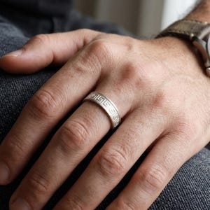 May include: A silver-toned band ring featuring a continuous engraved Greek key pattern around the exterior surface. The metal has a matte finish, and the geometric design consists of repeating interlocking square motifs that wrap around the ring.