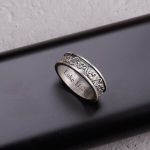 May include: A silver ring with an ornate, swirling pattern around the band. The text "Luke 11:28" is engraved on the inside of the ring. The ring is displayed on a dark surface.
