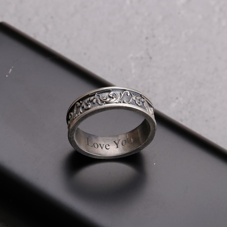 May include: A silver ring with an ornate, scrolling design around the band. The ring has the words "Love You" engraved on the inside. The ring is set against a dark background.