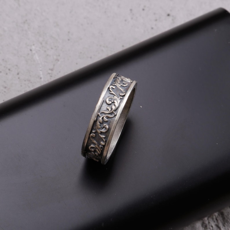 May include: A silver ring with an ornate, engraved design. The ring features a repeating pattern of swirling floral and scrollwork. The metal has a slightly darkened, antique finish, and the ring is set against a dark background.