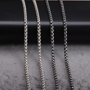 May include: Close-up of four silver-toned box chain necklaces. The chains are displayed vertically against a dark background and a textured gray surface. The chains vary slightly in color and finish, showcasing different styles.