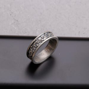 May include: A silver-colored ring with an ornate, swirling floral pattern around the band. The ring has a slightly oxidized finish, giving it a vintage or antique appearance. The design is detailed and raised, creating a textured surface.