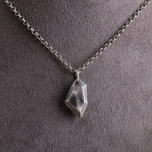 Könnte beinhalten: Halskette mit silbernem Anhänger, der ein geometrisches, facettiertes Edelsteindesign zeigt. Der Anhänger hat eine dunkle, strukturierte Oberfläche mit einem kleinen, kreisförmigen Muster. Die Halskette hat eine filigrane Gliederkette.