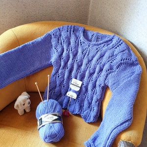 K&ouml;nnte beinhalten: Ein lila gestrickter Pullover mit einem Zopfmuster, der auf einem gelben Sessel liegt. Eine Kn&auml;uel lila Wolle mit dem Etikett "Alize" liegt auf dem Sessel neben dem Pullover.