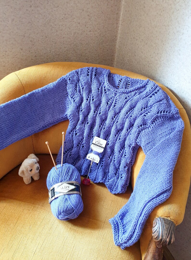 K&ouml;nnte beinhalten: Ein lila gestrickter Pullover mit einem Zopfmuster, der auf einem gelben Sessel liegt. Eine Kn&auml;uel lila Wolle mit dem Etikett "Alize" liegt auf dem Sessel neben dem Pullover.