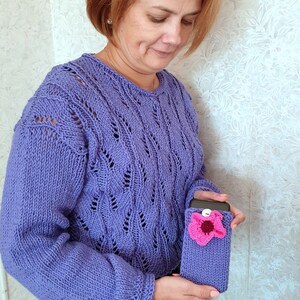 K&ouml;nnte beinhalten: Eine Frau tr&auml;gt einen lila gestrickten Pullover mit einer geh&auml;kelten Handyh&uuml;lle mit einem rosa Blumendesign.