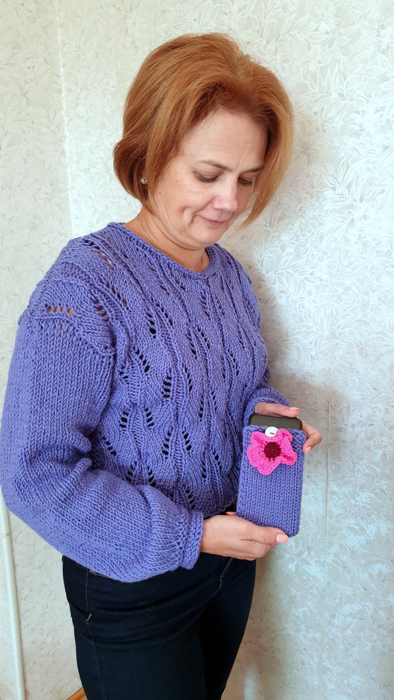 K&ouml;nnte beinhalten: Eine Frau tr&auml;gt einen lila gestrickten Pullover mit einer geh&auml;kelten Handyh&uuml;lle mit einem rosa Blumendesign.