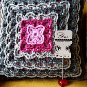 Op de afbeelding: Een grijs en roze gehaakt kussen met een vierkant patroon. Het kussen heeft een label met de tekst "Elena Fashion Handmade in Lithuania".