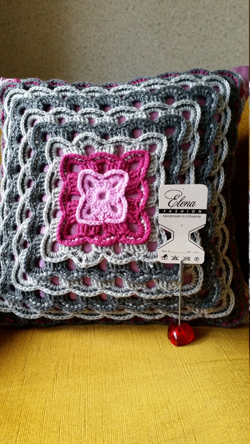 Op de afbeelding: Een grijs en roze gehaakt kussen met een vierkant patroon. Het kussen heeft een label met de tekst "Elena Fashion Handmade in Lithuania".