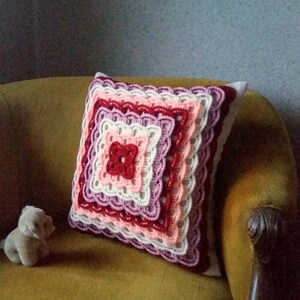 May include: A crocheted cushion with a square design in shades of pink, red, and white. The cushion is sitting on a brown armchair.