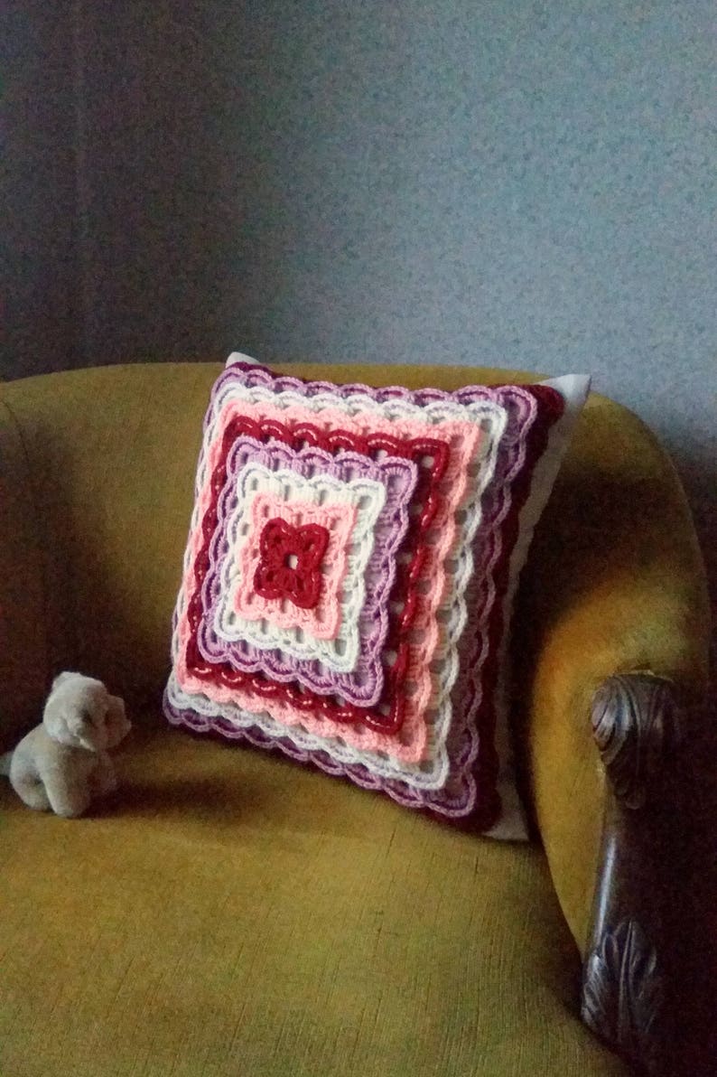 May include: A crocheted cushion with a square design in shades of pink, red, and white. The cushion is sitting on a brown armchair.