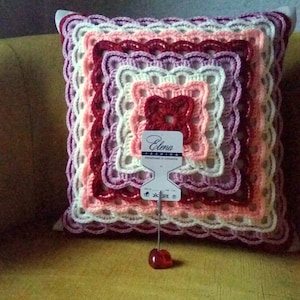 May include: A crocheted cushion with a square design in shades of pink, purple and red. The cushion has a white tag with the text "Elena Fashion Handmade in Lithuania".