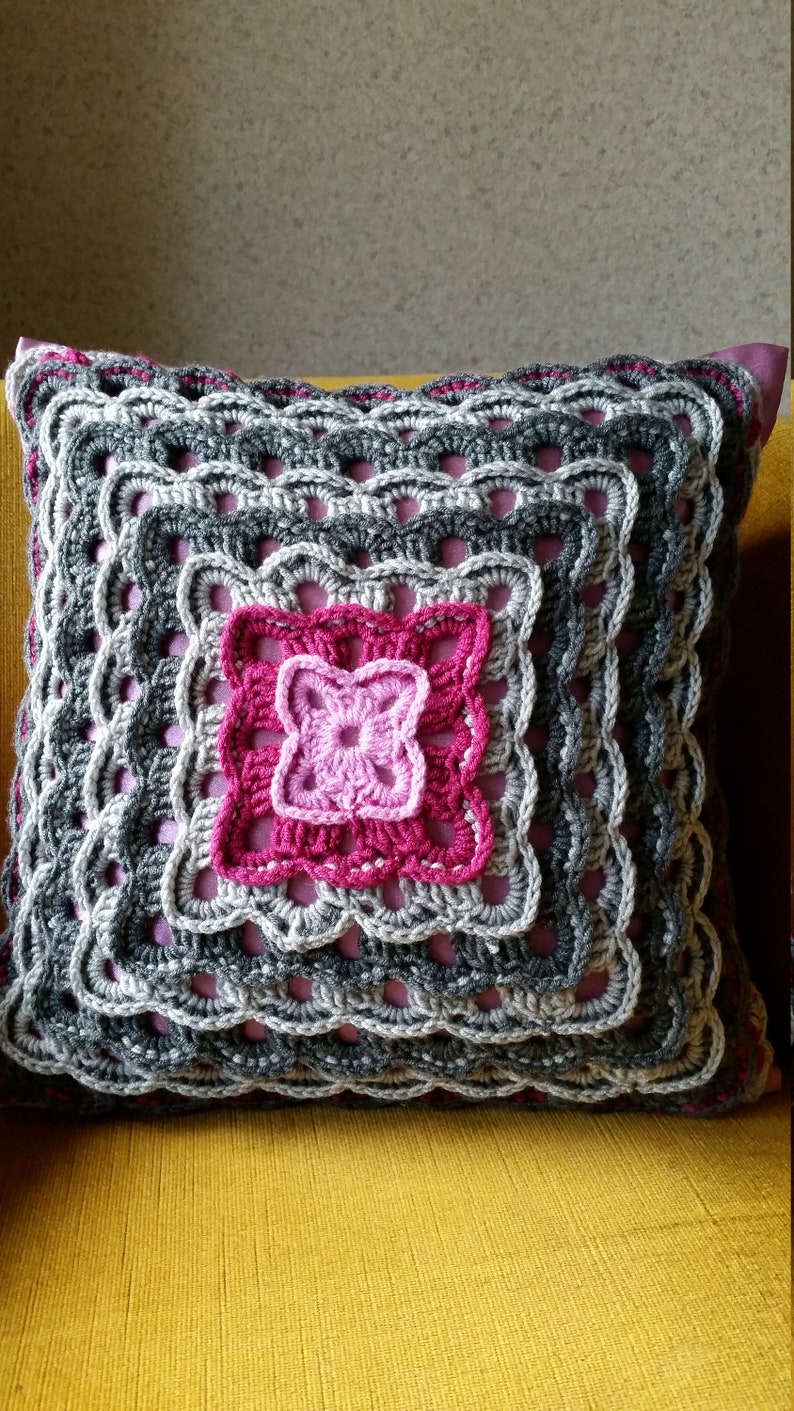 Op de afbeelding: Een gehaakt kussen met een geometrisch patroon in grijs en roze. Het kussen is vierkant en heeft een roze achterkant.