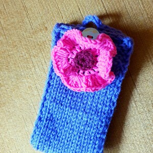 K&ouml;nnte beinhalten: Eine blaue gestrickte Handyh&uuml;lle mit einer rosa geh&auml;kelten Blume und einem silbernen Knopf.