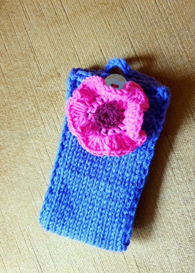 K&ouml;nnte beinhalten: Eine blaue gestrickte Handyh&uuml;lle mit einer rosa geh&auml;kelten Blume und einem silbernen Knopf.