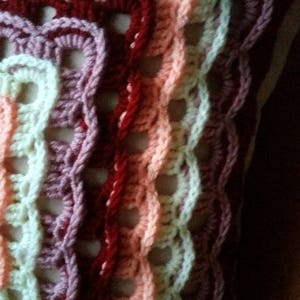 May include: A crocheted cushion featuring a multi-coloured design. The cushion is crafted using a range of pink, red, and white yarn shades. The crocheted pattern creates a textured and intricate design.