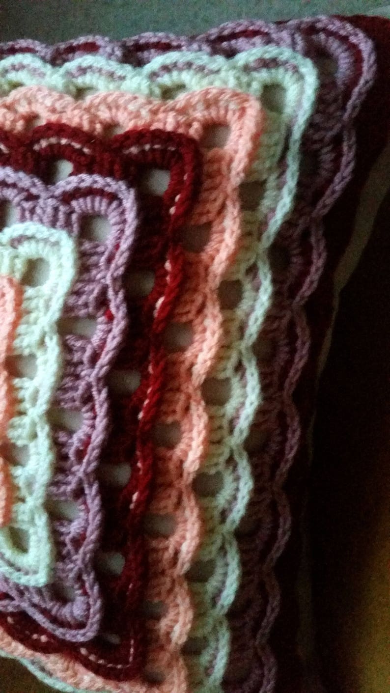 May include: A crocheted cushion featuring a multi-coloured design. The cushion is crafted using a range of pink, red, and white yarn shades. The crocheted pattern creates a textured and intricate design.