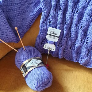 K&ouml;nnte beinhalten: Eine lila Garnrolle mit der Aufschrift "Alize LanaGold Classic" neben einem lila gestrickten Pullover mit einem Etikett mit der Aufschrift "Elena Fashion Handmade in Lithuania".