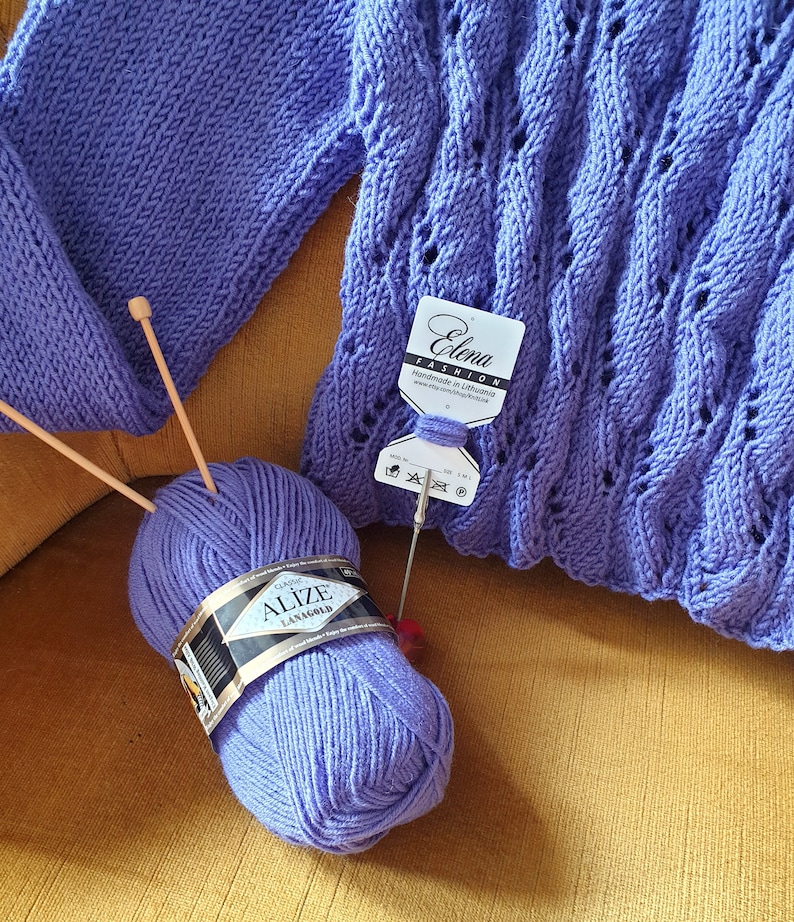 K&ouml;nnte beinhalten: Eine lila Garnrolle mit der Aufschrift "Alize LanaGold Classic" neben einem lila gestrickten Pullover mit einem Etikett mit der Aufschrift "Elena Fashion Handmade in Lithuania".