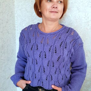 K&ouml;nnte beinhalten: Ein lila gestrickter Pullover mit einem Spitzenmuster. Der Pullover hat einen runden Ausschnitt und lange &Auml;rmel.