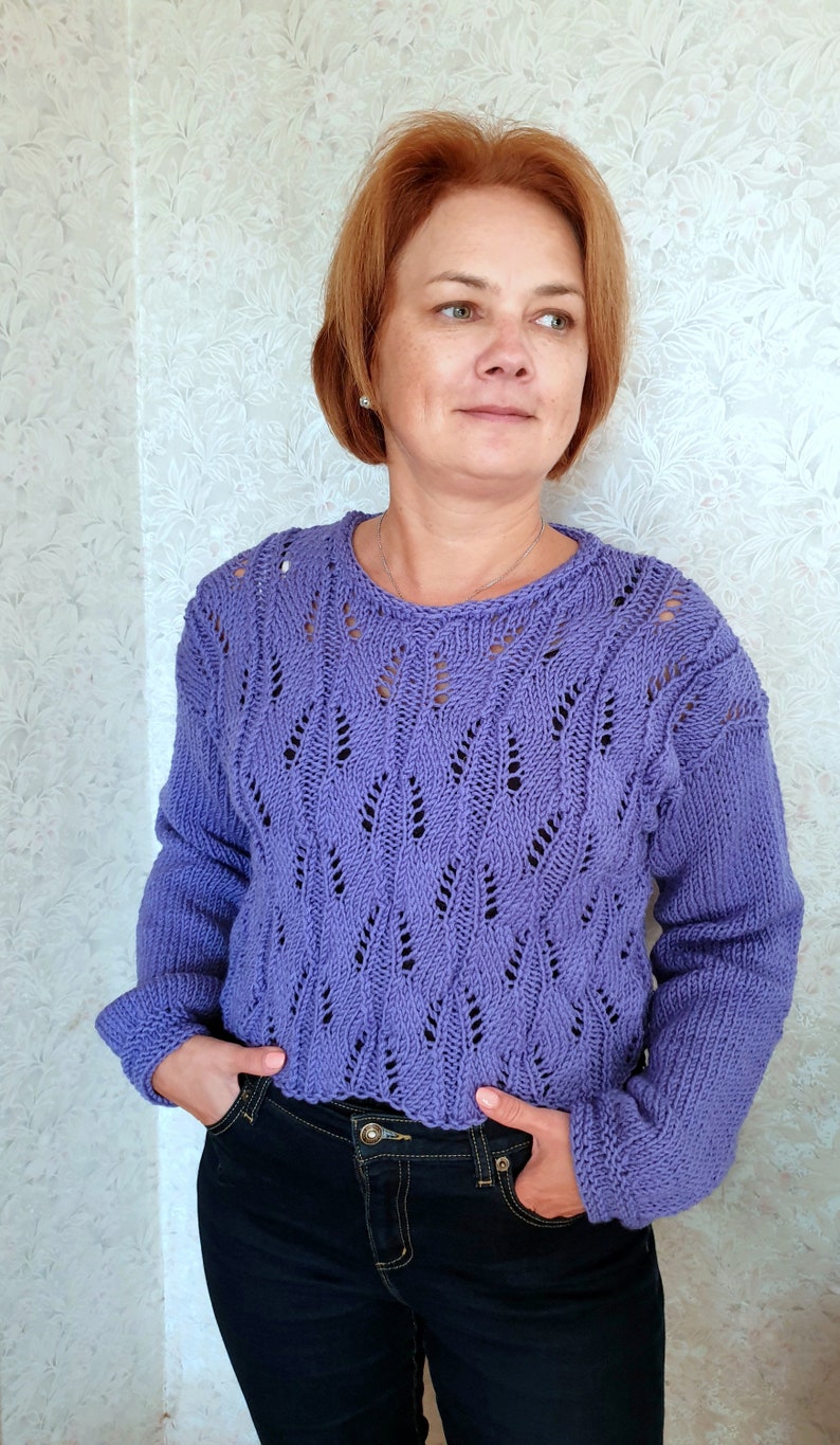 K&ouml;nnte beinhalten: Ein lila gestrickter Pullover mit einem Spitzenmuster. Der Pullover hat einen runden Ausschnitt und lange &Auml;rmel.