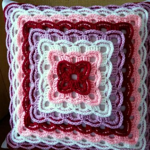May include: A crocheted cushion with a square design in shades of pink, white, and red. The cushion has a scalloped edge and is made with a variety of stitches.