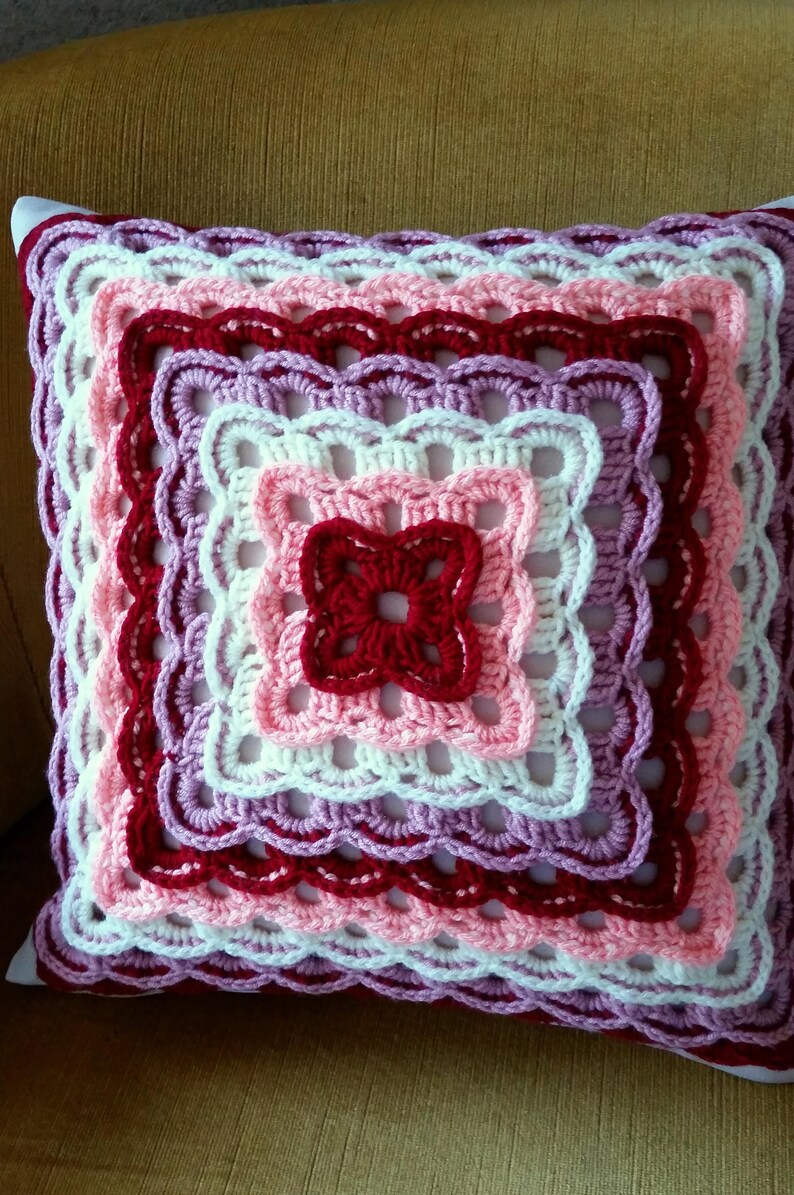 May include: A crocheted cushion with a square design in shades of pink, white, and red. The cushion has a scalloped edge and is made with a variety of stitches.