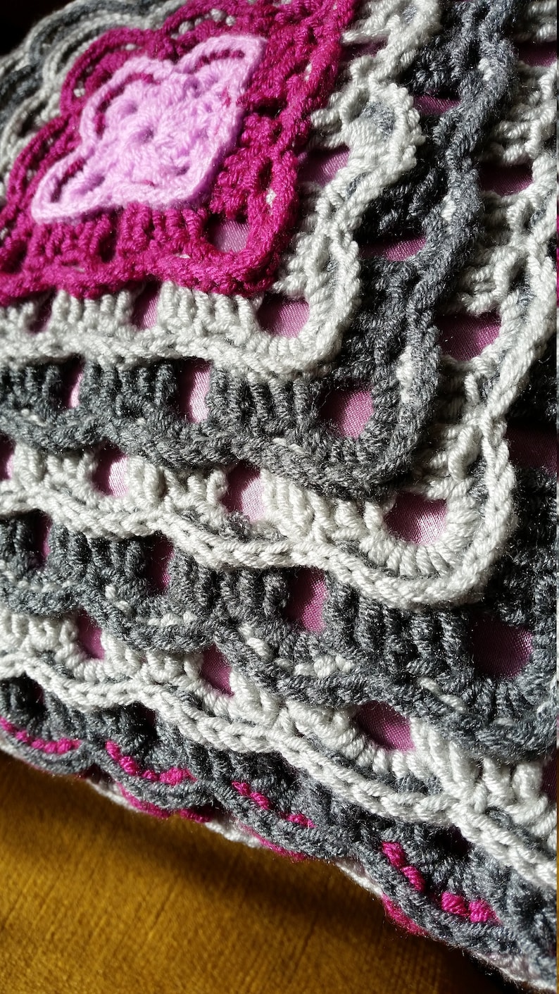 Op de afbeelding: Een close-up van een gehaakt kussen met een roze en grijs patroon. Het kussen is gemaakt met verschillende steken en texturen, wat een uniek en interessant ontwerp cre&euml;ert.