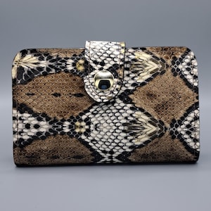 Könnte beinhalten: Eine rechteckige Geldbörse mit Schlangenmuster in Braun-, Schwarz- und Weißtönen. Die Geldbörse hat einen Verschluss mit einem goldfarbenen Metallakzent. Das Gesamtdesign ist ein Reptilienhautmuster.