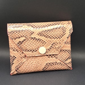 Pode incluir: Uma pequena carteira quadrada com um padrão de pele de cobra em tons de castanho e bege. A carteira tem um fecho de aba preso por um botão redondo de cor clara. As bordas são aparadas com um material rosa.