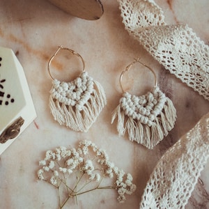 Può includere: Un paio di orecchini in macramè fatti a mano con cerchi dorati e frange bianche. Gli orecchini sono su una superficie di marmo con un nastro di pizzo bianco e una piccola scatola di legno.