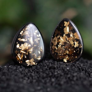 May include: Two teardrop-shaped black plugs with gold leaf flakes suspended within. The plugs are displayed on a bed of small, dark pebbles. The plugs have a glossy, polished finish, and the gold leaf adds a touch of luxury.