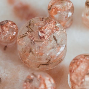 May include: A collection of clear, resin ear plugs with embedded copper-colored flakes and small plant matter. The plugs vary in size and shape, with a soft, diffused light. The overall aesthetic is delicate and natural, suggesting handmade jewelry.
