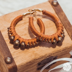 May include: A pair of large hoop earrings made of brown yarn and gold beads. The earrings are on a wooden surface.