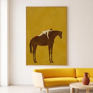 May include: A framed rectangular wall art print featuring a dark brown horse standing in profile against a solid mustard yellow background. A figure in a white long-sleeved top and red socks is lying horizontally across the horse's back.