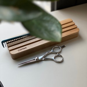 Op de afbeelding: Een houten kam- en schaarhouder met het woord "COMBO" gegraveerd op de voorkant. Een zilveren schaar met een zwarte handgreep ligt op een wit oppervlak.
