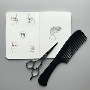 May include: An open notebook featuring black ink technical diagrams of hair sectioning and a profile sketch of a hairstyle, accompanied by a pair of silver metal hair cutting shears and a black plastic hair comb with fine and wide teeth.